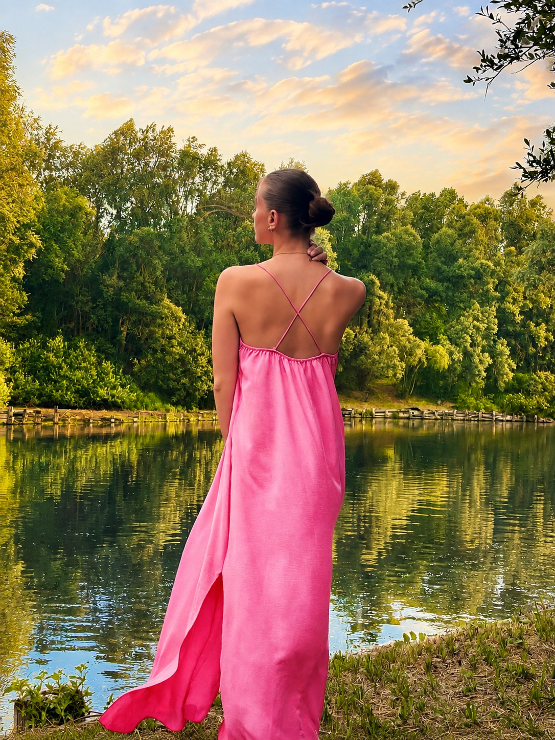 Femme de dos en robe rose satinée face à un lac au coucher du soleil